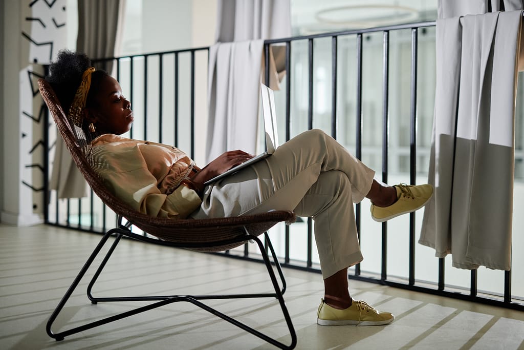 A black woman wearing cream clothing relaxing in a chair on a balcony with a laptop on her lap